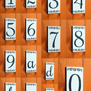 Puede incluir: Azulejos de cerámica rectangulares blancos con números y letras negros del 0 al 9, a, b, c, d y una barra inclinada. Cada azulejo tiene un borde floral en azul, naranja y verde. Los azulejos están montados sobre una superficie de madera naranja.