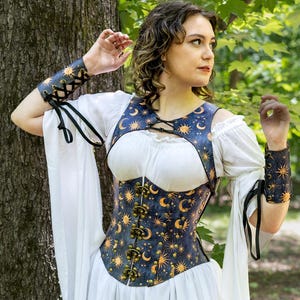 May include: A white dress with long sleeves and a layered skirt. The dress is paired with a navy blue corset adorned with gold moons and stars. Matching arm cuffs complete the celestial-themed ensemble. Brown boots are visible.