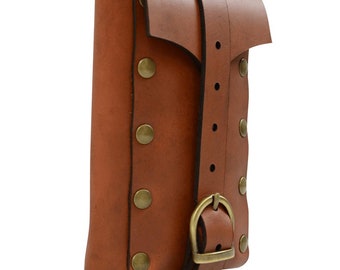 Op de afbeelding: Een bruin leren tasje met een klepsluiting en een gesp. Het tasje is vastgezet met een riem en heeft messingkleurige klinknagels. Het ontwerp doet denken aan middeleeuwse of renaissance-accessoires.