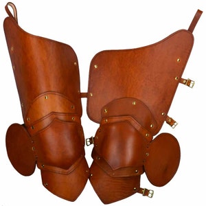 May include: A pair of brown leather knee pads with metal accents and buckles. The pads are designed to protect the knees during activities such as horseback riding or medieval reenactment.