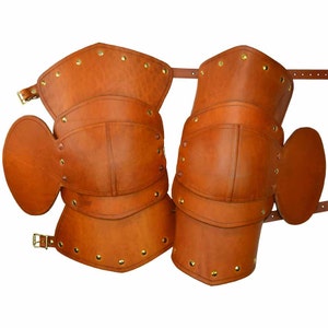 May include: A pair of brown leather knee pads with brass rivets and straps. The knee pads are designed to protect the knees from injury.