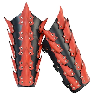 Op de afbeelding: Een paar rode en zwarte leren armbeschermers met een draken schubben ontwerp. De beschermers zijn versierd met zilveren studs.