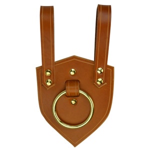May include: A brown leather shield-shaped sheath with a gold-colored ring and rivets. Two brown leather straps extend from the top. The sheath is designed to hold a sword or other bladed weapon.