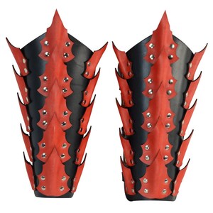 Op de afbeelding: Een paar rode en zwarte leren armbeschermers met een stekelig ontwerp. De beschermers zijn versierd met zilveren studs.