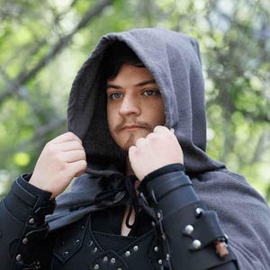 May include: A person wearing a gray hooded cloak with a black leather vest and arm guards. The hood is pulled up over their head, and they are looking directly at the camera.