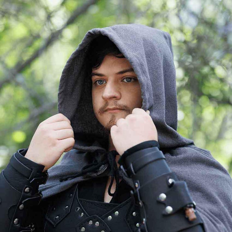 May include: A person wearing a gray hooded cloak with a black leather vest and arm guards. The hood is pulled up over their head, and they are looking directly at the camera.