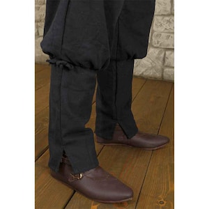 K&ouml;nnte beinhalten: Schwarze mittelalterliche Hose mit gerafftem Oberbein und geteiltem Unterbein. Die Hose wird mit braunen Lederschuhen mit Schnallenverschluss getragen. Das Outfit befindet sich auf einem Holzboden.