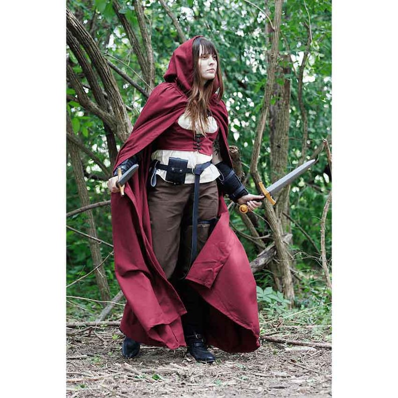 May include: A woman wearing a red hooded cloak and brown pants holds a sword in a forest setting.