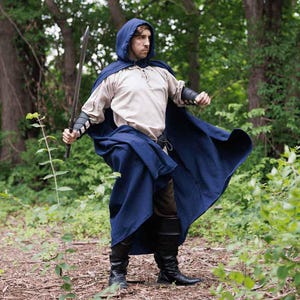 May include: A person wearing a blue hooded cloak with a sword in their hand. The cloak is flowing in the wind and the person is standing in a forest.
