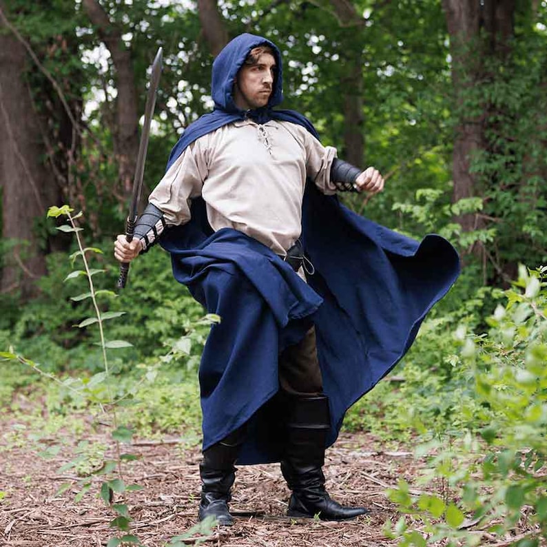 May include: A person wearing a blue hooded cloak with a sword in their hand. The cloak is flowing in the wind and the person is standing in a forest.