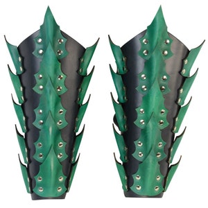 Op de afbeelding: Een paar groene en zwarte leren armbeschermers met een stekelig ontwerp. De beschermers zijn gemaakt van leer en hebben een reeks stekels die langs de lengte van de arm lopen. De stekels zijn gemaakt van metaal en zijn met klinknagels aan het leer bevestigd.