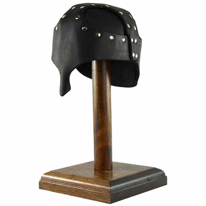 Puede incluir: Casco de cuero negro con tachuelas met&aacute;licas, exhibido en un soporte de madera. El casco tiene una parte superior puntiaguda y una solapa que cubre la parte posterior del cuello.