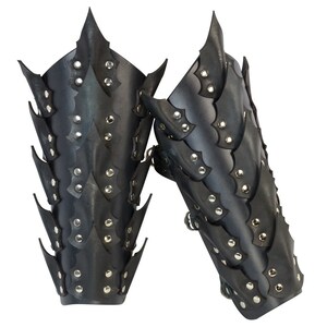 Op de afbeelding: Een paar zwarte leren armbeschermers met een stekelig, schubbig ontwerp. De beschermers zijn versierd met zilveren studs en hebben een sluiting met een band.