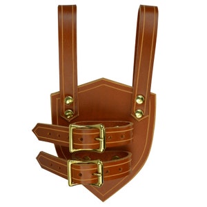 May include: A brown leather sword scabbard with two straps and buckles. The scabbard is shaped like a shield and has a decorative stitching detail.