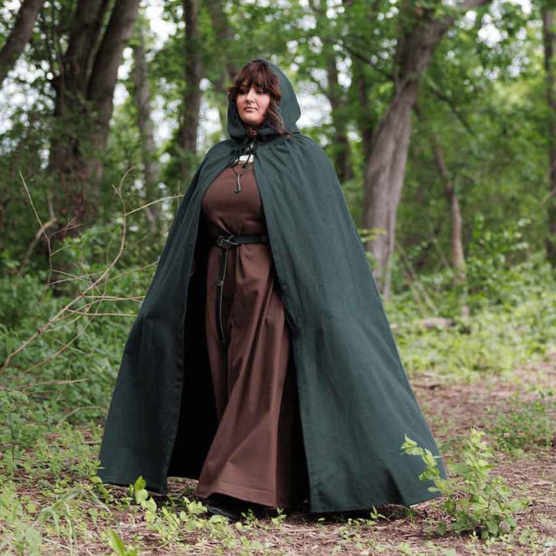May include: A woman wearing a long, dark green hooded cloak with a brown dress underneath. The cloak is made of a soft, flowing fabric and has a simple design. The woman is standing in a forest setting.