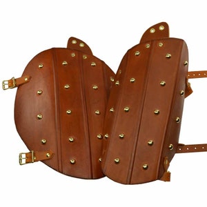 May include: A pair of brown leather leg guards with brass rivets and buckles. The guards are designed to protect the shins and knees.