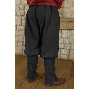 K&ouml;nnte beinhalten: Schwarze mittelalterliche Hose mit geraffter Taille und lockerer Passform. Die Hose hat einen breiten, weiten oberen Teil, der sich zu einem eng anliegenden Unterschenkel mit einem Schlitz am Kn&ouml;chel verj&uuml;ngt. Die Hose ist aus dunklem Stoff gefertigt und eignet sich f&uuml;r historische Nachstellungen oder Kost&uuml;me.