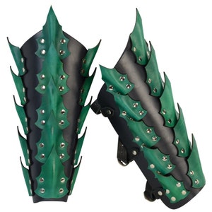 Op de afbeelding: Een paar groene en zwarte leren armbeschermers met spikes. De beschermers zijn gemaakt van leer en hebben een groene en zwarte kleurstelling. De spikes zijn van metaal en zijn bevestigd aan het leer.