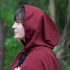 May include: A woman wearing a deep red hooded cloak. The cloak is made of a soft, textured fabric and has a simple design.