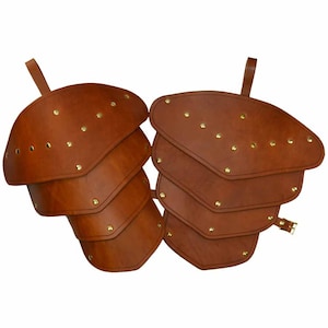May include: A pair of brown leather shoulder armor with brass accents. The armor is made of multiple overlapping plates and has a strap for fastening.