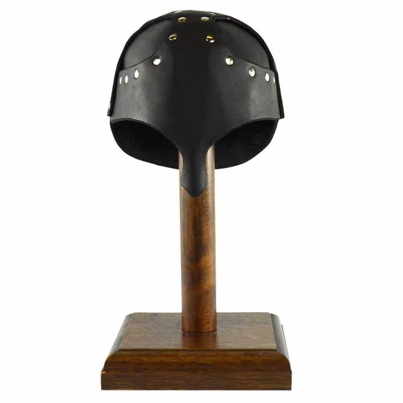 Puede incluir: Casco de cuero negro con remaches de lat&oacute;n, exhibido en un soporte de madera.