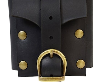 Puede incluir: Brazalete de cuero negro con solapa y cierre de hebilla. El brazalete presenta remaches de color latón y una hebilla a juego. El diseño es sencillo y funcional.