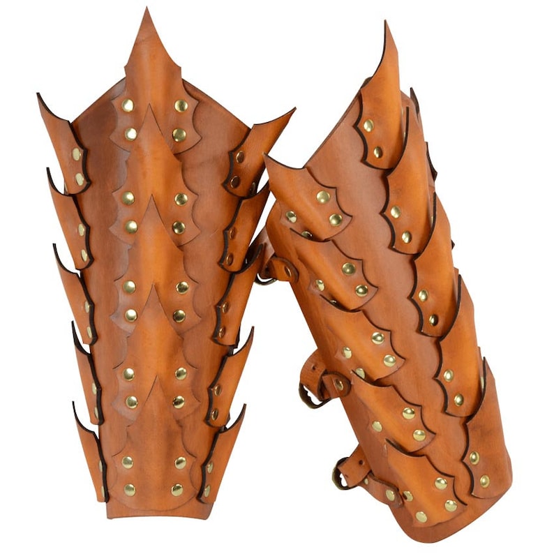 Op de afbeelding: Een paar bruine leren armbeschermers met een schubbenpatroon en gouden klinknagels. De beschermers zijn ontworpen om de onderarmen te beschermen en hebben een puntig, stekelig uiterlijk.