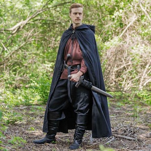 May include: A person wearing a black hooded cloak with a brown shirt and black pants. They are holding a sword in their right hand.