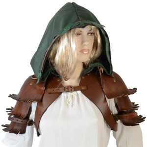 May include: Brown leather shoulder armor with a green hooded cloak. The armor has a fringed design and is secured with straps and buckles. The hood has a green leather trim and is lined with a soft material.