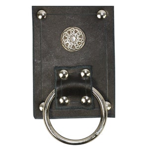 May include: A black leather belt loop with a large silver ring. The loop is adorned with silver rivets and a decorative silver button with a Celtic knot design. The leather has a textured appearance.