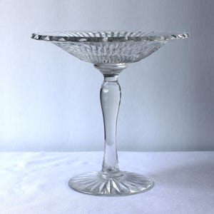 May include: A clear glass pedestal bowl featuring a wide, shallow basin with a scalloped, fluted rim and a radiating sunburst pattern. The bowl is supported by a slender, smooth stem that connects to a circular base with matching radial detailing.