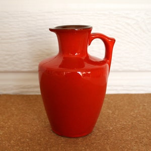 May include: A red ceramic pitcher with a brown rim and a handle. The pitcher is sitting on a cork surface.