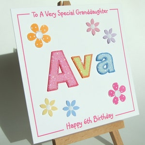 May include: A white card with pink, yellow, blue, and orange glitter letters spelling "Ava" with a pink border. The card has a message that says "To A Very Special Granddaughter" and "Happy 6th Birthday" with colorful flower embellishments.