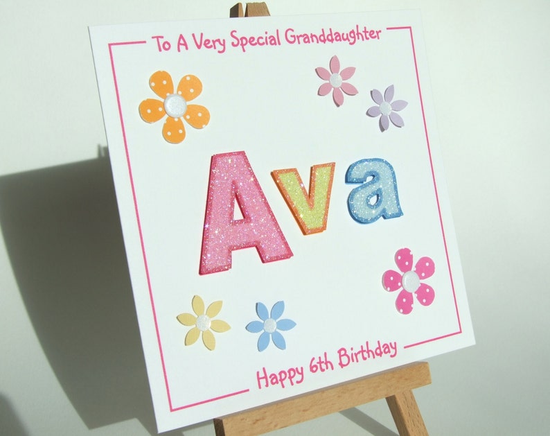May include: A white card with pink, yellow, blue, and orange glitter letters spelling "Ava" with a pink border. The card has a message that says "To A Very Special Granddaughter" and "Happy 6th Birthday" with colorful flower embellishments.