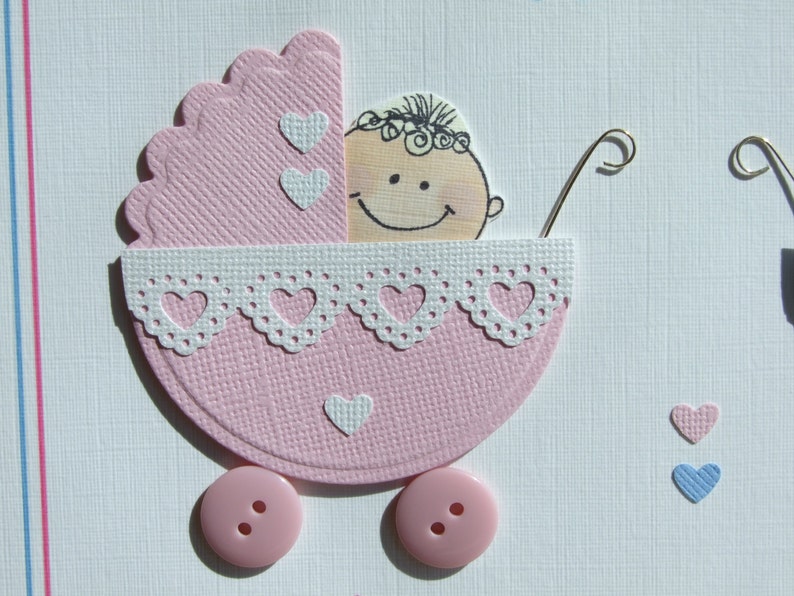 May include: A pink baby carriage with a white lace trim and a smiling baby face inside. The carriage has two pink buttons for wheels and two white hearts on the side.
