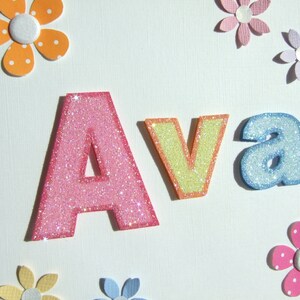 May include: A white card with the name "Ava" spelled out in glittery letters. The letters are pink, yellow, and blue. The card is decorated with colorful paper flowers.