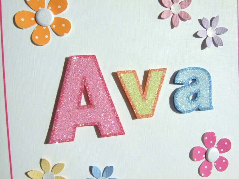 May include: A white card with the name "Ava" spelled out in glittery letters. The letters are pink, yellow, and blue. The card is decorated with colorful paper flowers.