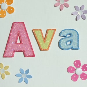 May include: A white card with the name "Ava" spelled out in pink, yellow, and blue glitter letters. The letters are surrounded by colorful paper flowers.
