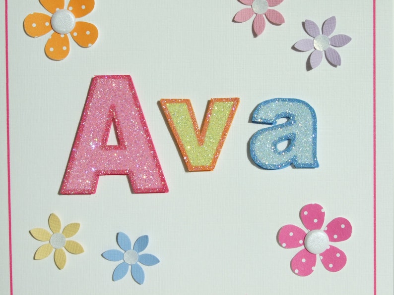 May include: A white card with the name "Ava" spelled out in pink, yellow, and blue glitter letters. The letters are surrounded by colorful paper flowers.