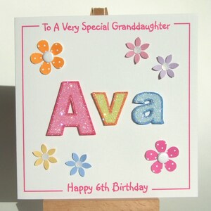 May include: A white card with pink trim and the words "To A Very Special Granddaughter" at the top. The name "Ava" is spelled out in large, glittery letters in pink, yellow, and blue. There are several colorful paper flowers around the name and the words "Happy 6th Birthday" at the bottom.