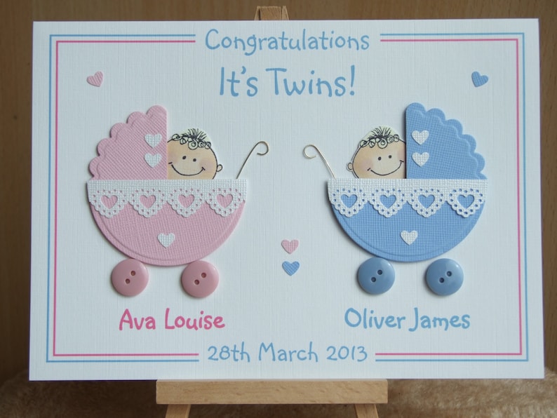 May include: A white card with a pink and blue baby carriage design. The card says "Congratulations It's Twins!" and includes the names "Ava Louise" and "Oliver James" with the date "28th March 2013".