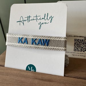Puede incluir: Una pulsera tejida gris y blanca con el texto bordado en azul "KA KAW" se muestra en una tarjeta blanca. La tarjeta tambi&eacute;n presenta las palabras "Authentically you" en escritura turquesa.
