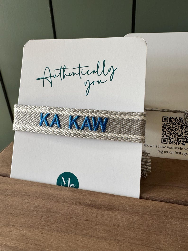 Puede incluir: Una pulsera tejida gris y blanca con el texto bordado en azul "KA KAW" se muestra en una tarjeta blanca. La tarjeta tambi&eacute;n presenta las palabras "Authentically you" en escritura turquesa.