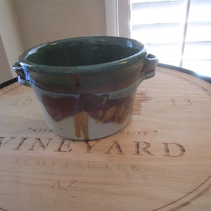 May include: A round ceramic bowl with two small side handles, featuring a dark green glazed rim and a mottled body with patches of deep red, brown, and cream colors. The bowl has a wide opening and a smooth, glossy finish on the upper section.
