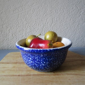 May include: A blue and white speckled ceramic bowl filled with fruit. The bowl is on a wooden cutting board. The fruit includes a red pepper, a green pear, and two yellow pears.
