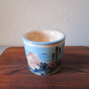 May include: A small, blue and white ceramic planter with a desert scene painted on the side. The scene includes a mountain, a cactus, and some small plants.