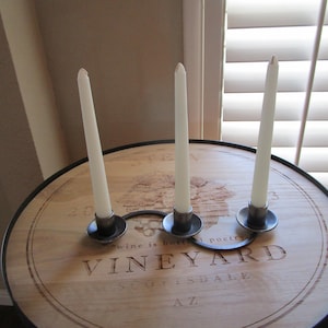 May include: Three white candles on metal candle holders on a wooden table with the text "Vineyard" and "Southside AZ" engraved on the surface.