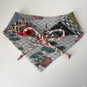 Puede incluir: Un pañuelo triangular para mascotas con tela de múltiples patrones y un lazo central. El diseño incluye motivos de olas grises, pinos verdes, detalles florales rojos y secciones geométricas negras. Un lazo de tela color crema está anudado en el centro.