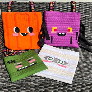 May include: Four quilted fabric bags featuring Halloween-themed designs: an orange pumpkin, a purple monster, a green Frankenstein-style creature, and a white mummy. Each bag displays a stylized face and includes black straps with pumpkin patterns.