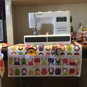 May include: A white Brother sewing machine sits on a quilted fabric cover featuring a colorful pattern of Russian nesting dolls. The cover includes multiple pockets holding sewing tools like a yellow rotary cutter and grey scissors.